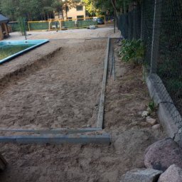 Huzar Maciej Możejko - Przygotowanie terenu pod budowę tarasu drewnianego z widocznymi obrzeżami i piaskiem, obok basen i ogrodzenie z siatki.