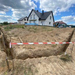 Huzar Maciej Możejko - Wykop pod fundamenty domu jednorodzinnego, zabezpieczony taśmą ostrzegawczą, z widocznymi szalunkami i pracującym robotnikiem w tle, na tle błękitnego nieba z chmurami.