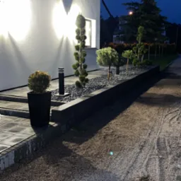 Nowoczesny ogród przydomowy z podświetloną elewacją, ozdobnymi krzewami w donicach i na rabacie wysypanej grysem, widok nocą.