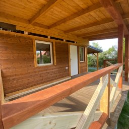 Huzar Maciej Możejko - Zadaszony taras z drewnianą konstrukcją, widoczne deski podłogowe, balustrada w trakcie montażu z elementami surowego drewna i pomalowanymi na brązowo, drewniana elewacja budynku w tle.