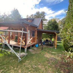 Huzar Maciej Możejko - Drewniany domek letniskowy z zadaszonym tarasem w trakcie budowy, widoczne rusztowanie i pracownik w zielonych spodniach, trawnik i drzewa w tle.