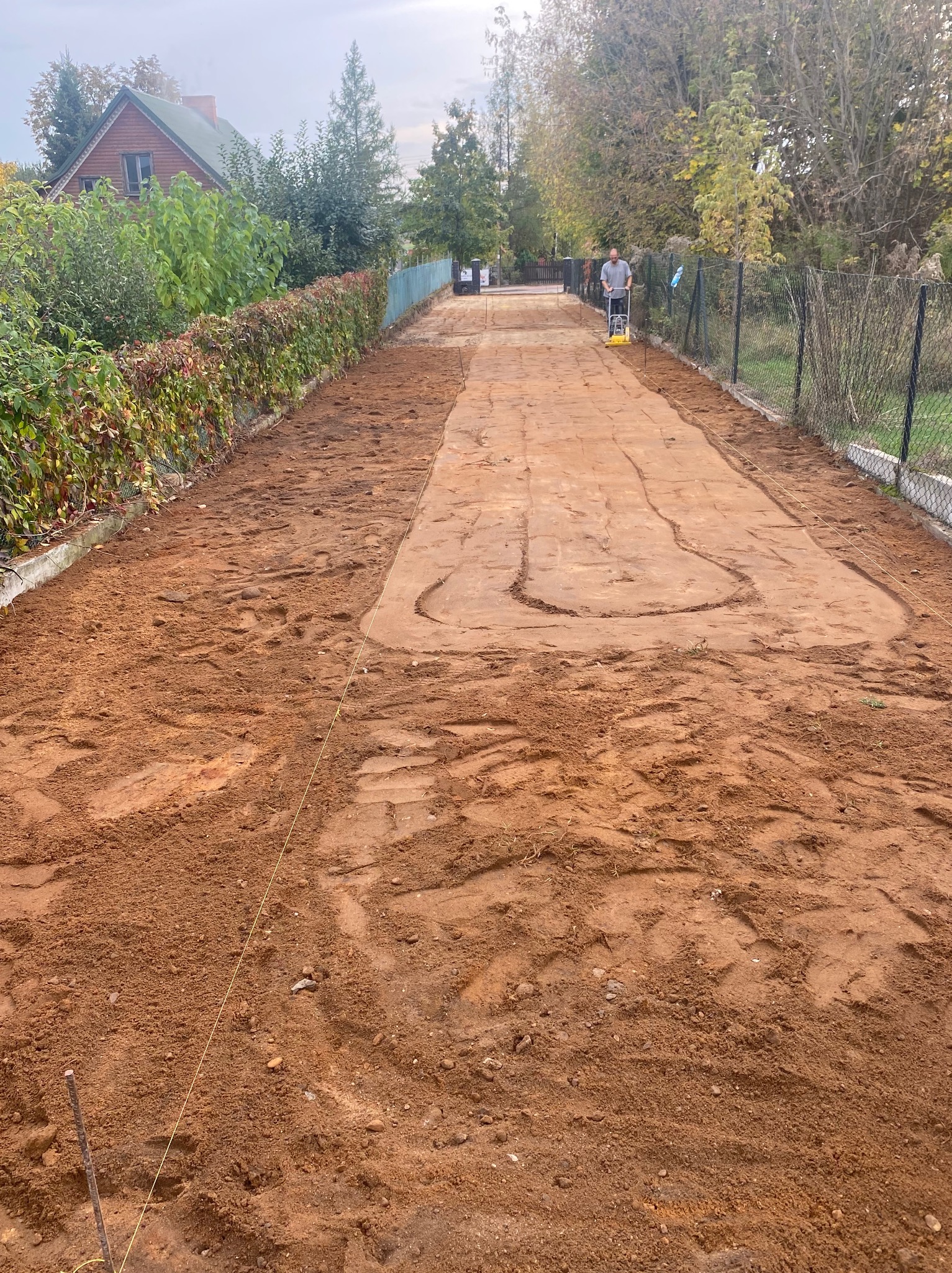 Przygotowanie podłoża pod ścieżkę z kostki brukowej: ubijanie ziemi zagęszczarką wzdłuż ogrodzenia, wytyczona linia, widoczne ślady opon.