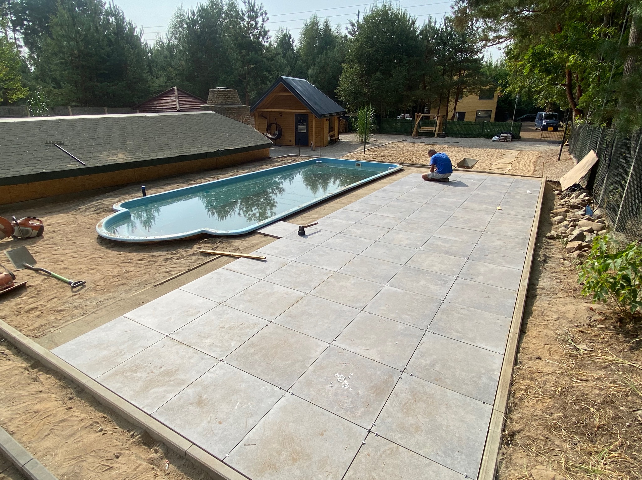 Układanie jasnoszarej kostki brukowej na piasku obok basenu w ogrodzie, widoczne krzyżyki dystansowe, narzędzia i pracownik w trakcie pracy.