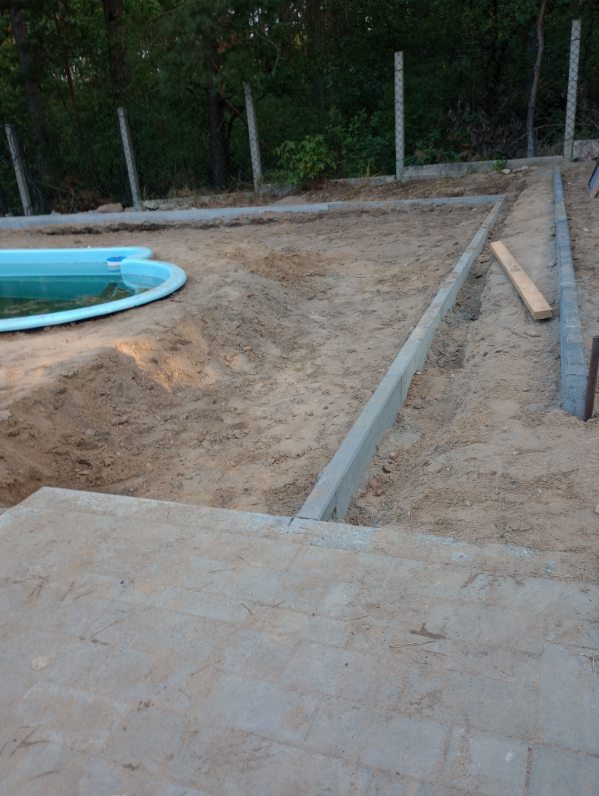 Przygotowanie podłoża pod kostkę brukową i basen ogrodowy, widoczne krawężniki i częściowo wykopany teren, ogrodzenie z siatki w tle.