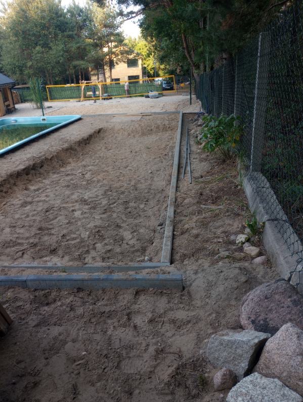 Przygotowanie terenu pod budowę tarasu drewnianego z widocznymi obrzeżami i piaskiem, obok basen i ogrodzenie z siatki.