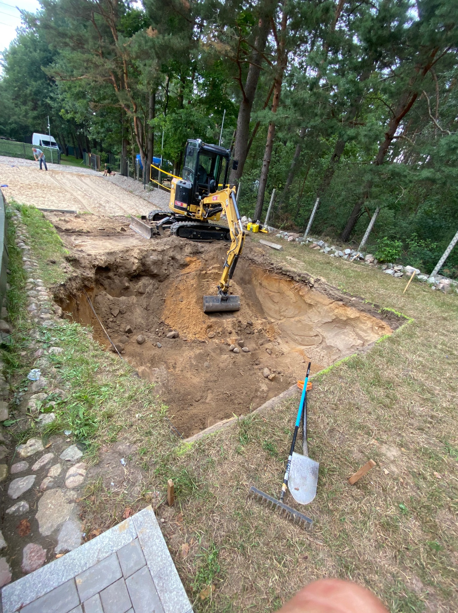 Żółta mini koparka w wykopie o nieregularnym kształcie na działce z drzewami w tle. Na trawie obok leżą łopata i grabie.