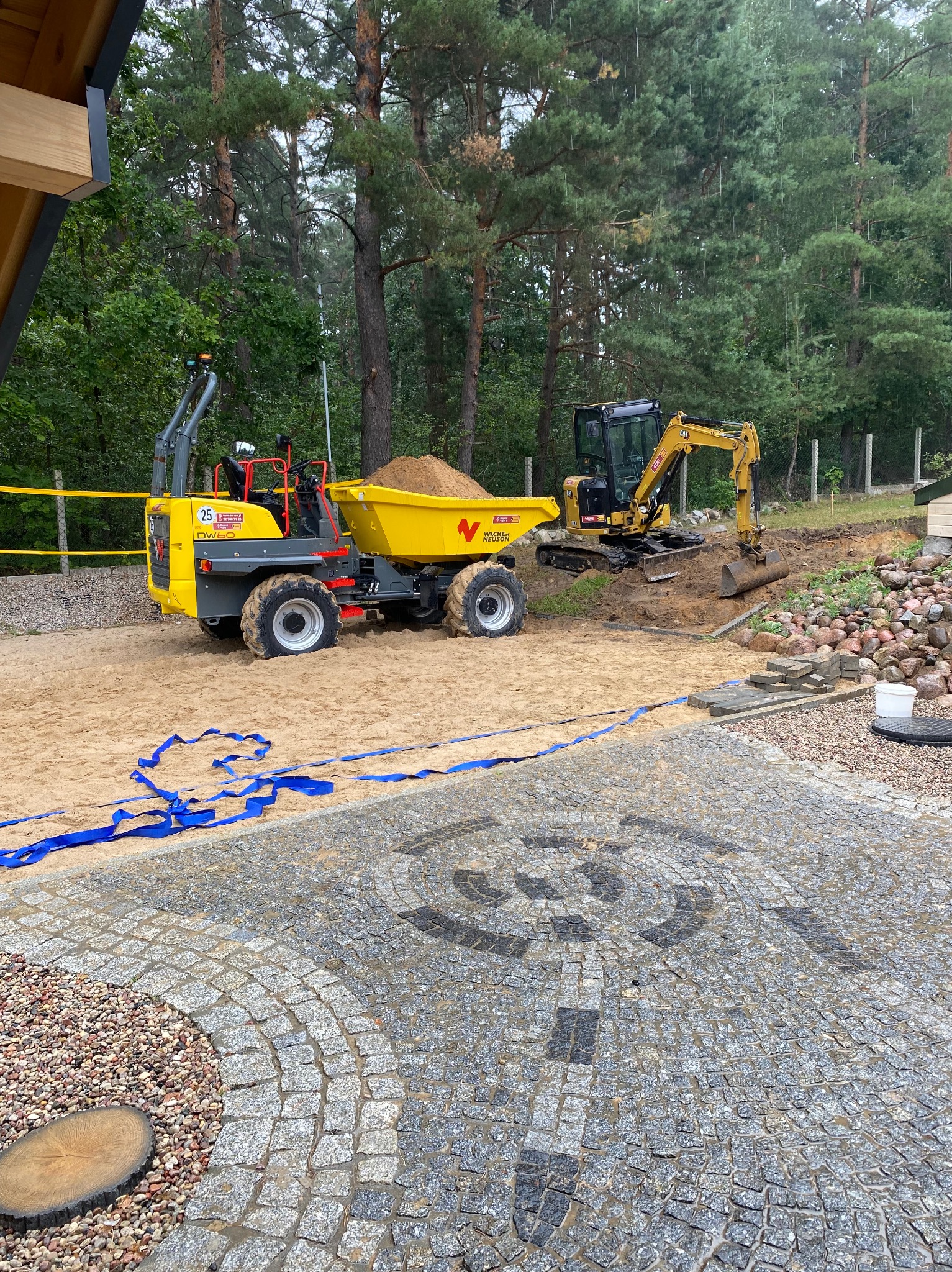 Żółty wozidło wypełnione piaskiem i mała koparka na placu budowy, widoczny fragment ułożonej kostki brukowej z ozdobnym wzorem, w tle drzewa.