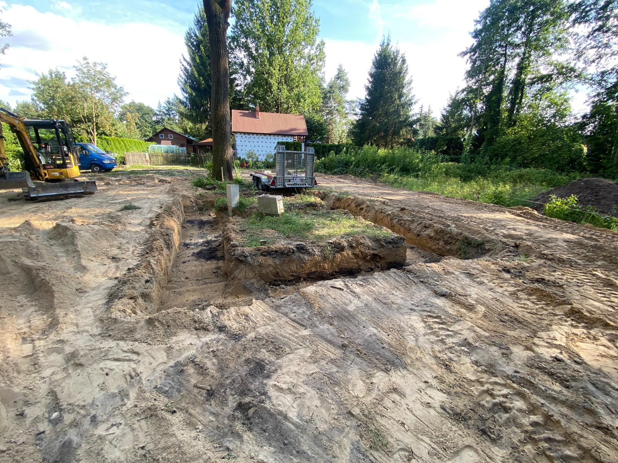 Wykop pod fundamenty z widocznym traktorem, przyczepką i elementami szalunku w ogrodzie z domem w tle.
