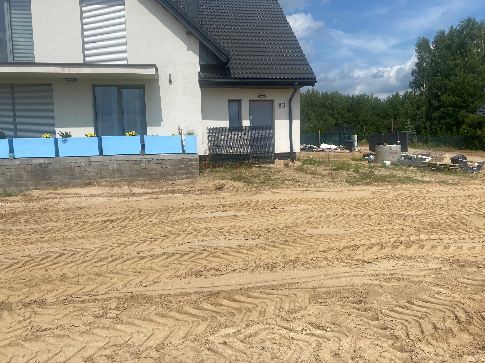 Nowoczesny dom jednorodzinny z numerem 83, widoczne ślady opon na piasku, przygotowania do prac ziemnych, w tle częściowo widoczny betonowy zbiornik i elementy ogrodzenia.