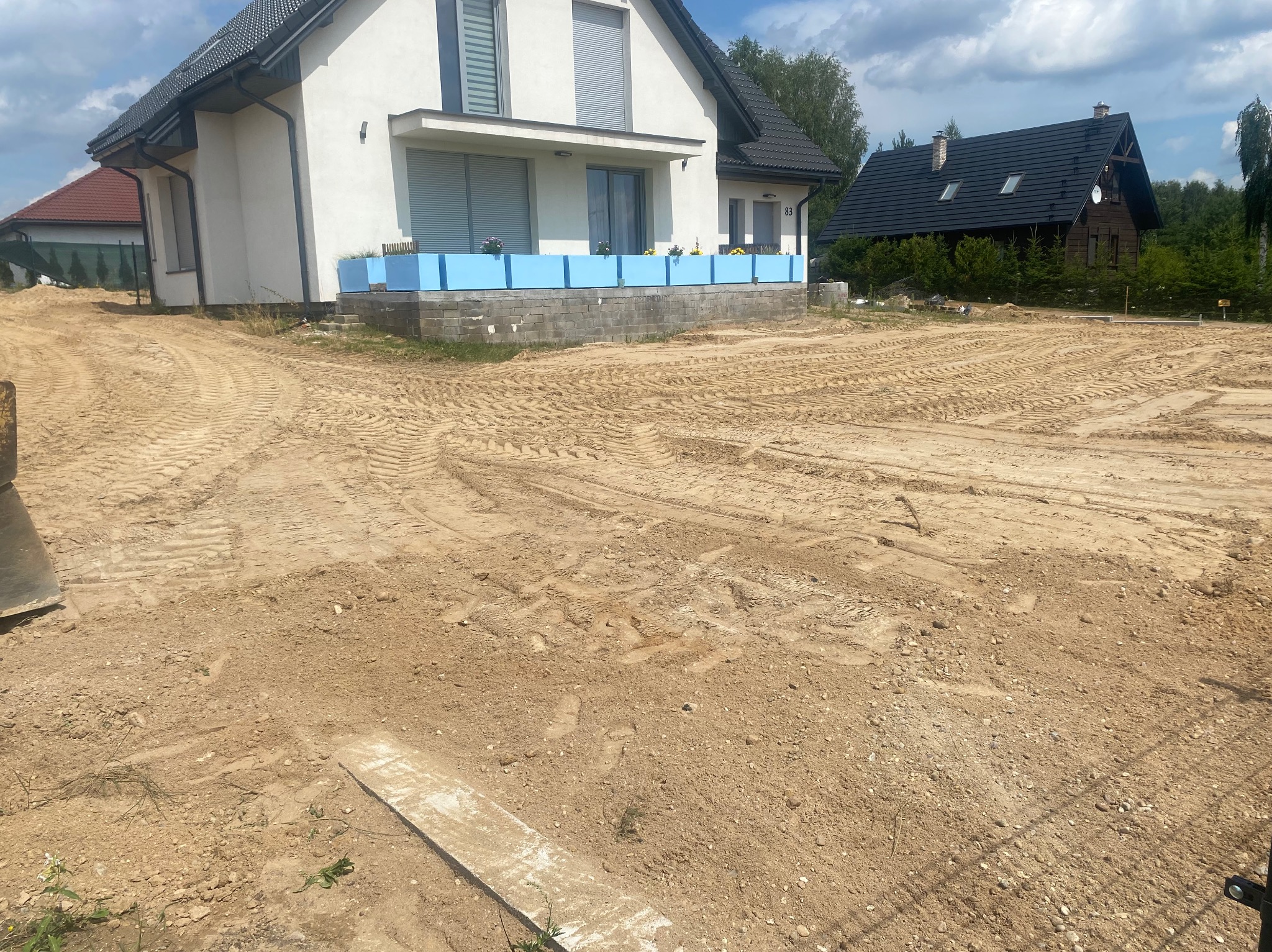 Przygotowany teren pod budowę z widocznym domem w tle, ślady opon i świeżo posadzonymi kwiatami w donicach na tarasie.