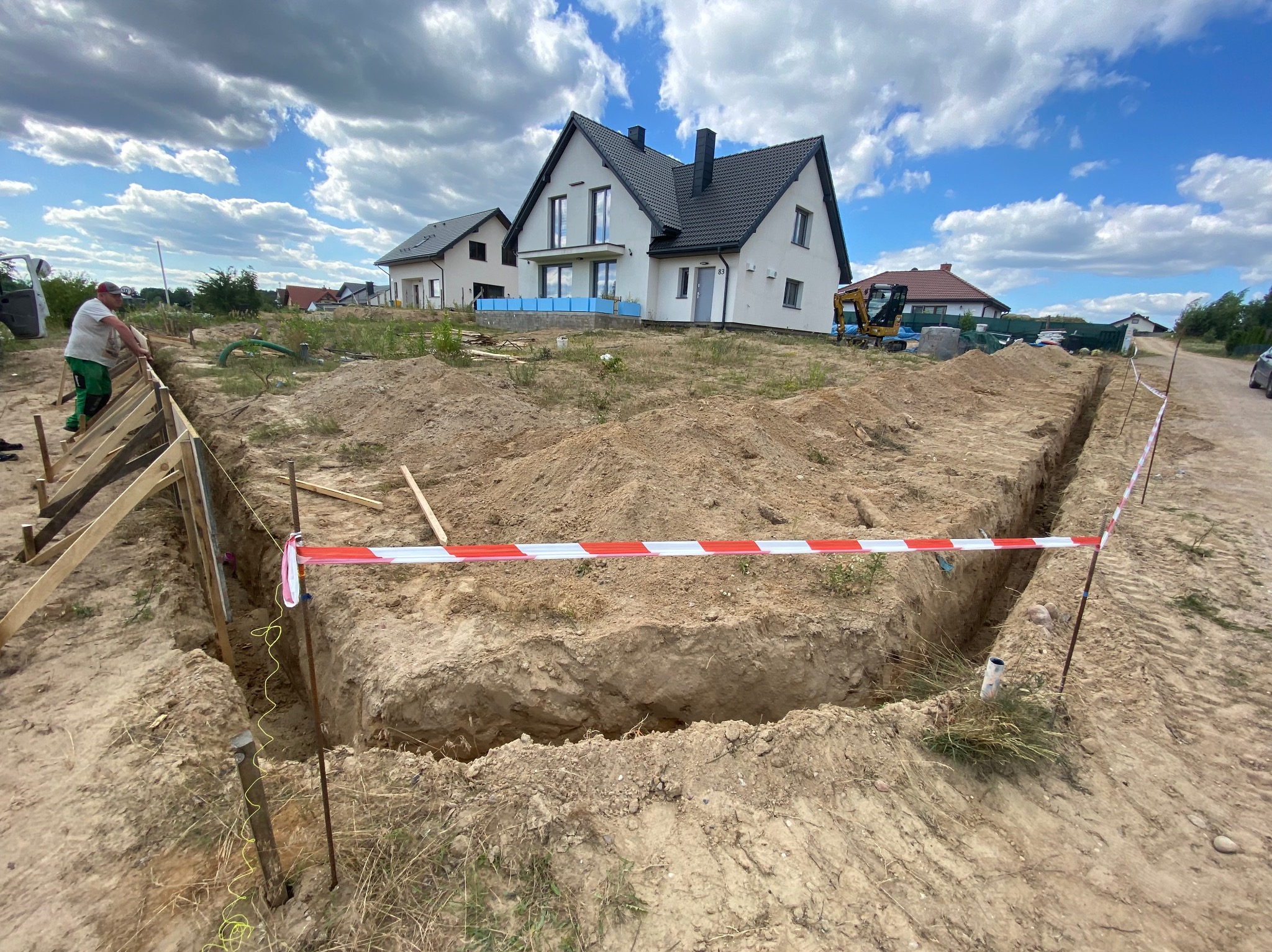 Wykop pod fundamenty domu jednorodzinnego, zabezpieczony taśmą ostrzegawczą, z widocznymi szalunkami i pracującym robotnikiem w tle, na tle błękitnego nieba z chmurami.