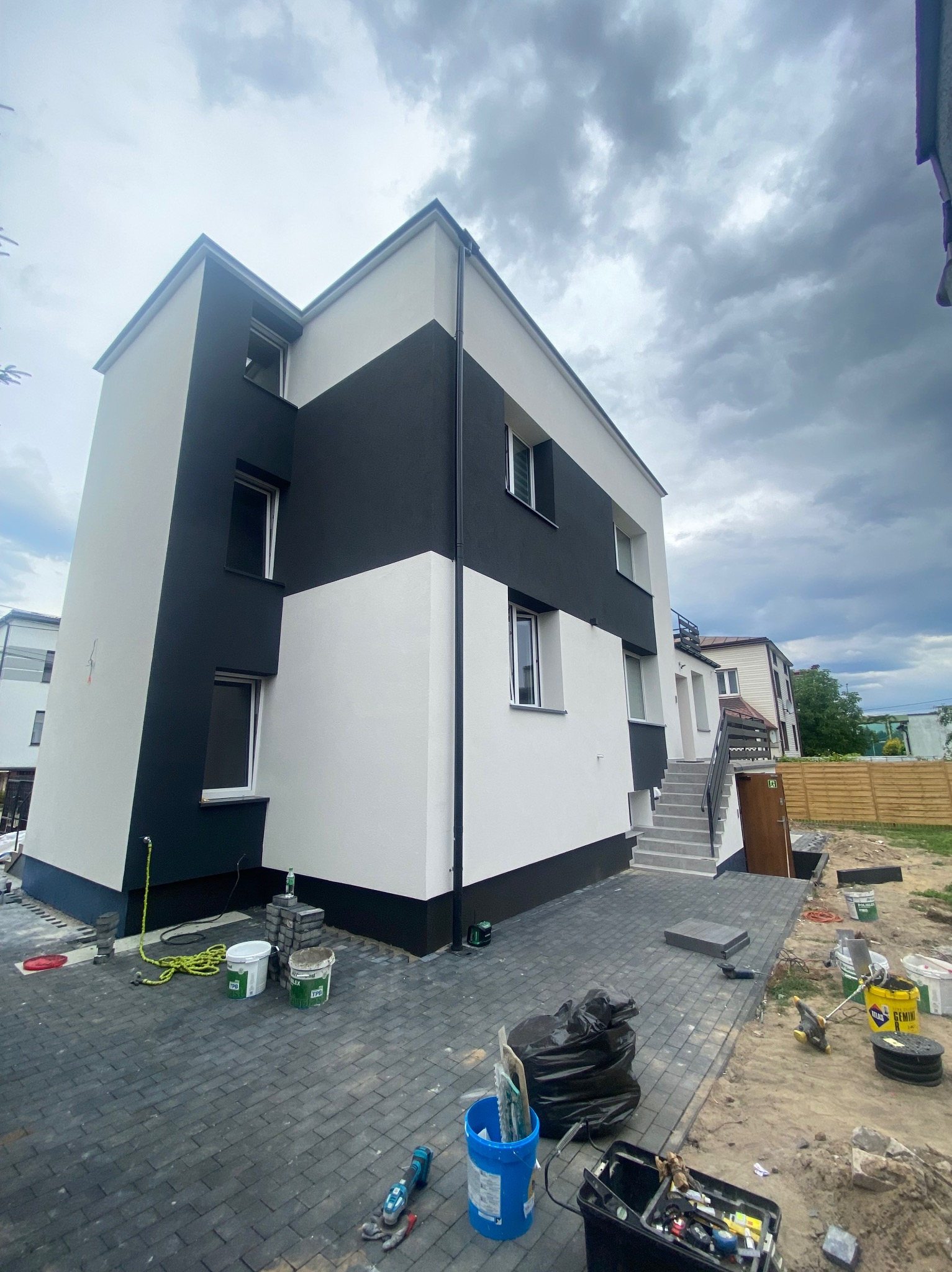 Dwukolorowa elewacja budynku w trakcie wykańczania, widoczne narzędzia i materiały budowlane na brukowanym podłożu, pochmurne niebo w tle.