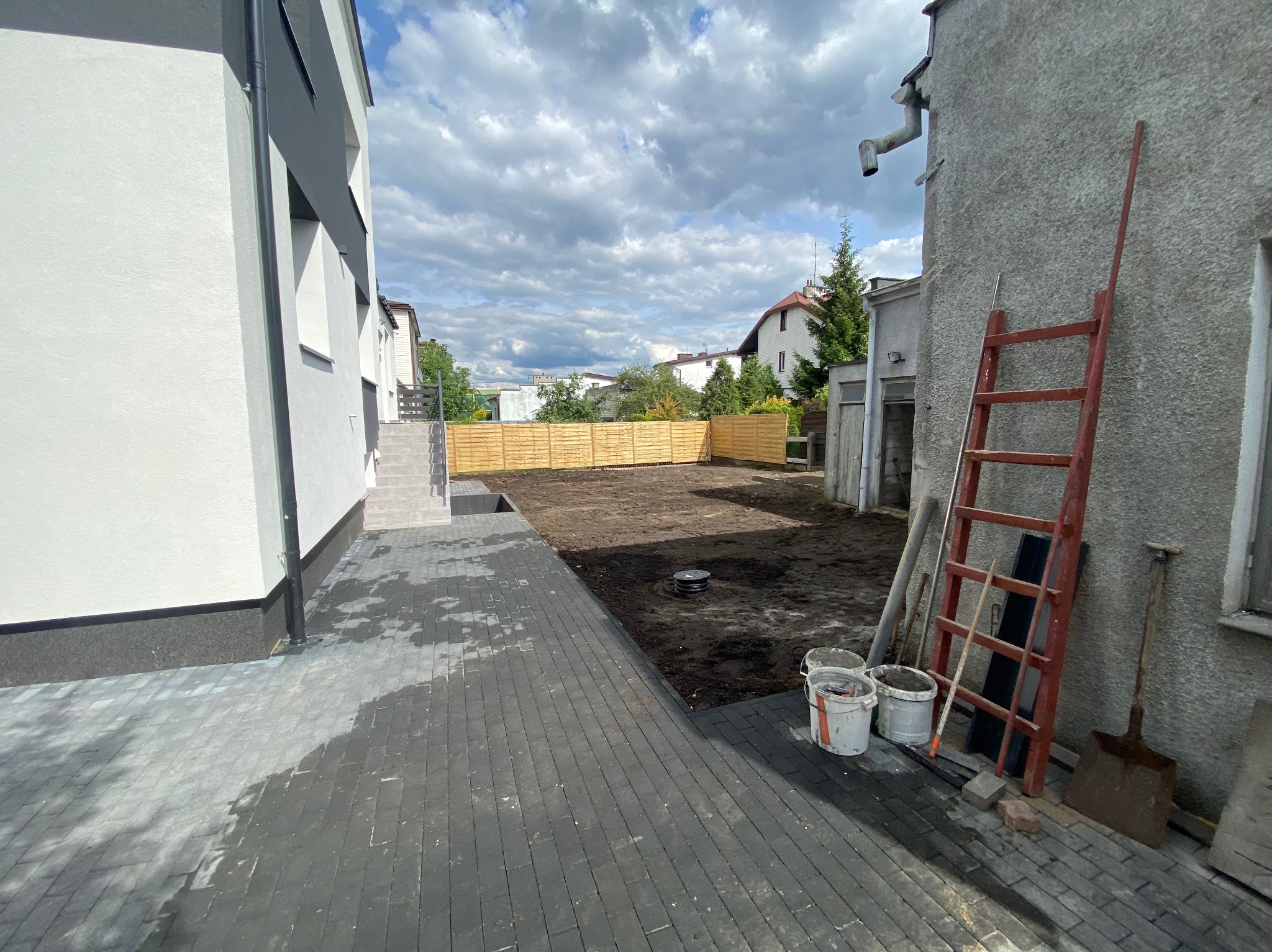 Świeżo ułożona, ciemnoszara kostka brukowa przed nowoczesnym domem, widoczny drewniany płot w tle, narzędzia i wiaderka malarskie oparte o starą ścianę budynku.