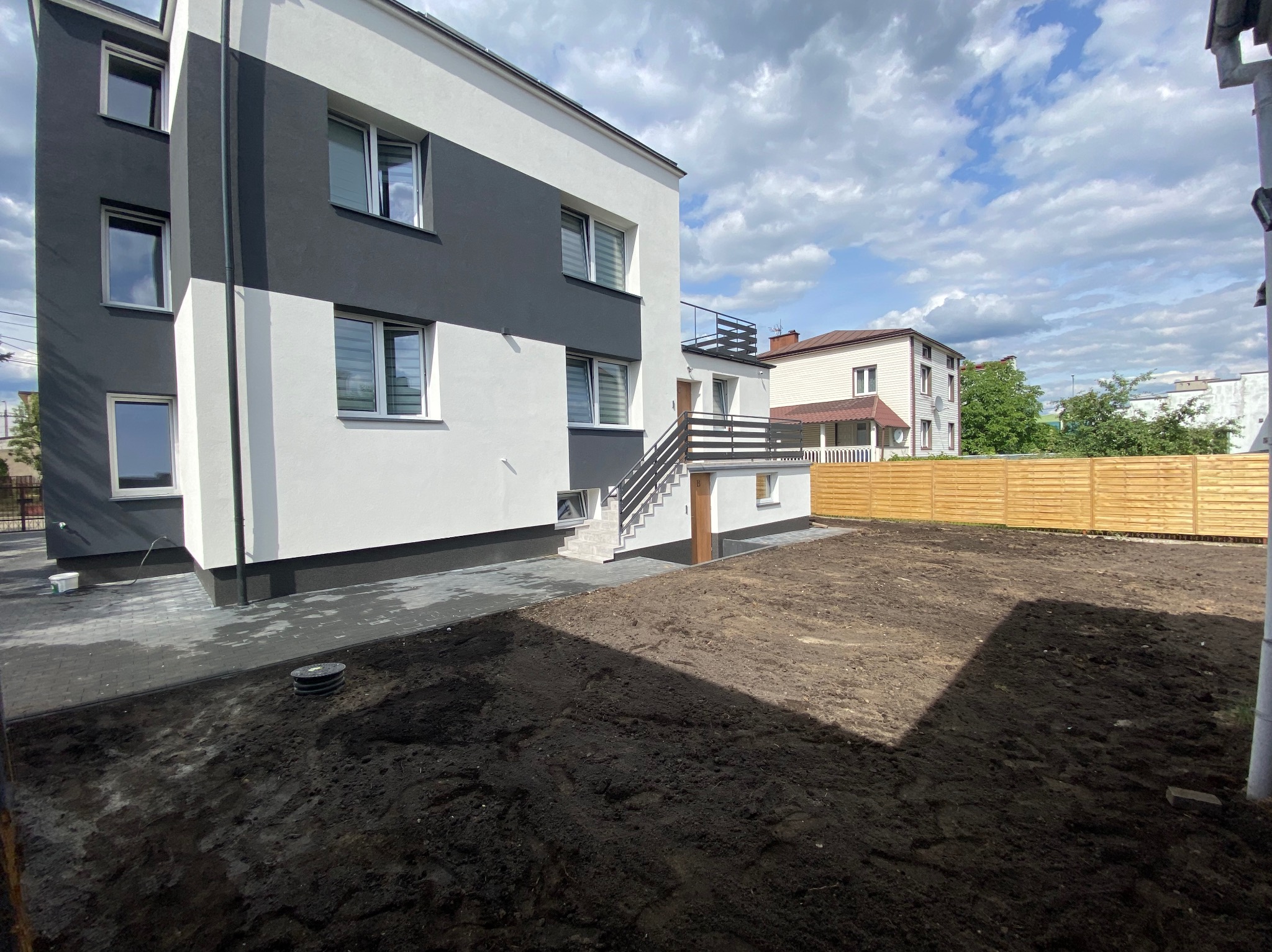 Nowoczesny budynek z elewacją w kolorach szarości i bieli, widoczne świeżo ułożone szare kostki brukowe i wyrównany teren przygotowany pod trawnik, w tle drewniane ogrodzenie.