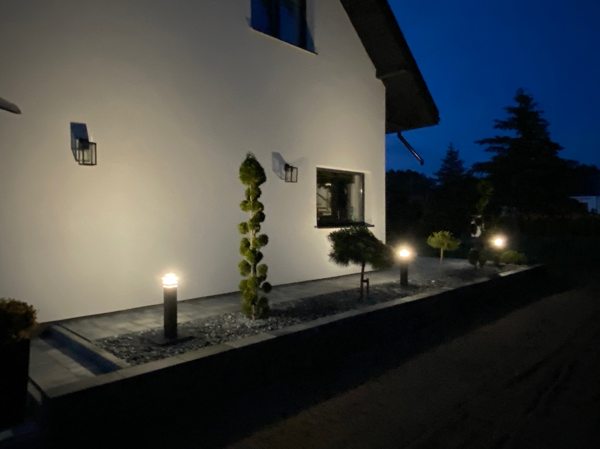 Oświetlona elewacja domu z ozdobnymi krzewami i ścieżką wyłożoną kostką brukową oraz lampami ogrodowymi, sfotografowana nocą.
