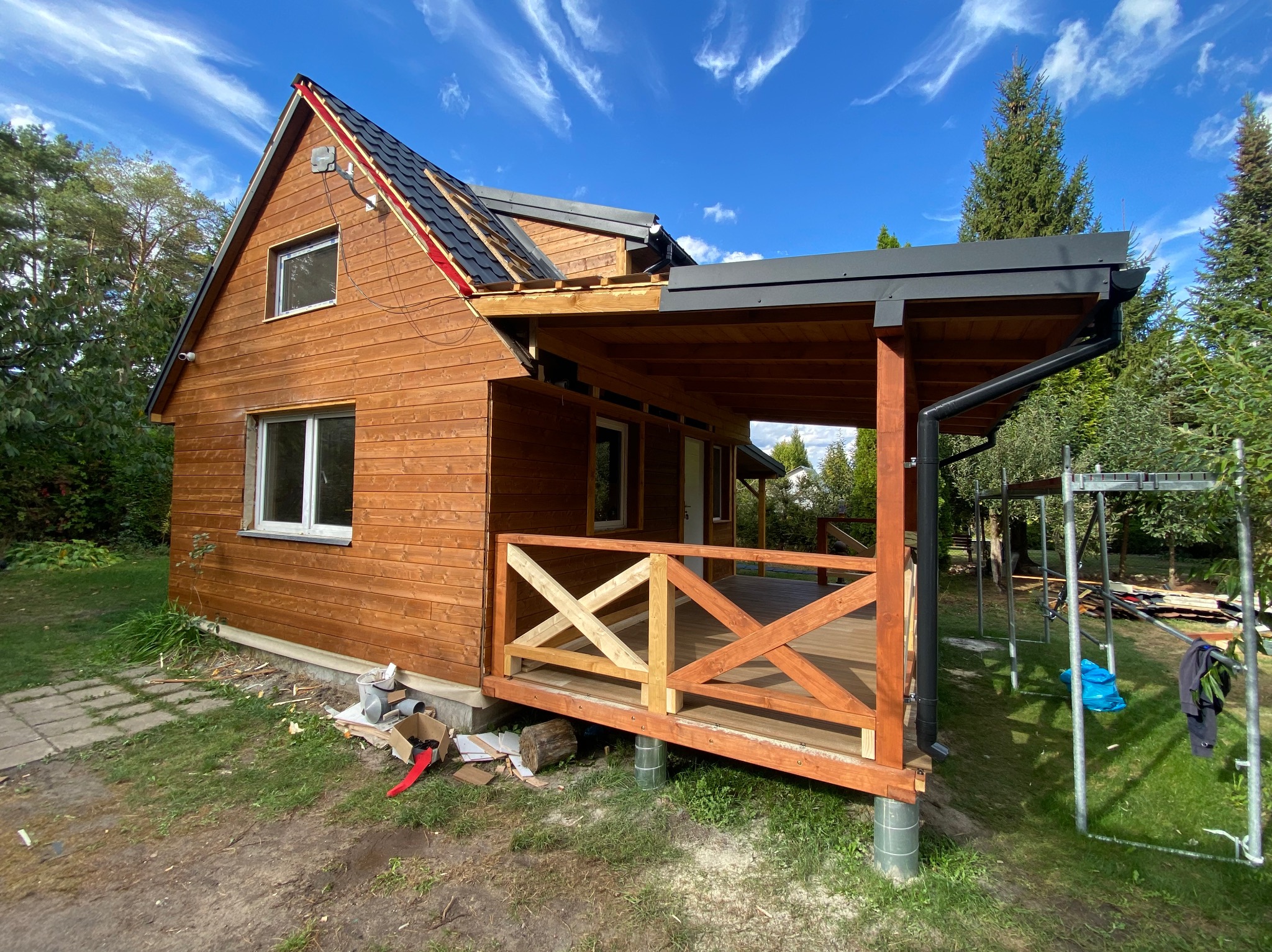 Drewniany dom w trakcie budowy: widoczna część elewacji z oknami, zadaszony taras z drewnianą balustradą w stylu krzyżowym, fragment dachu z częściowo ułożoną dachówką, rusztowania w tle.