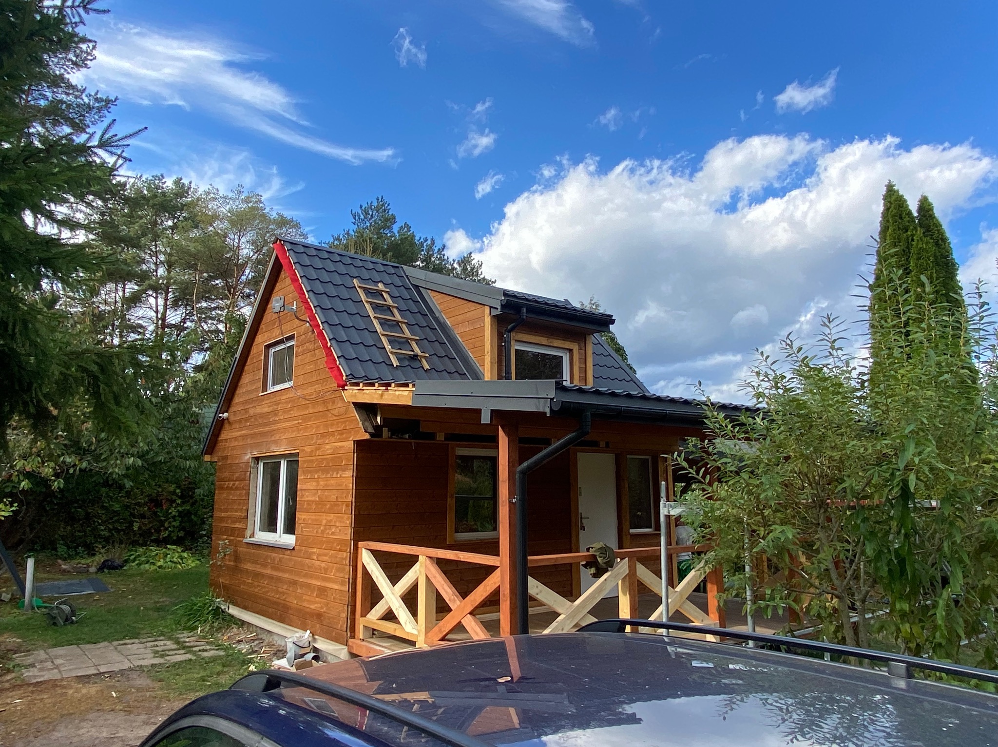 Drewniany dom jednorodzinny z czarną dachówką i drewnianą drabinką opartą o dach, w trakcie prac wykończeniowych elewacji i montażu drewnianej balustrady tarasu, na tle błękitnego nieba z obłokami.