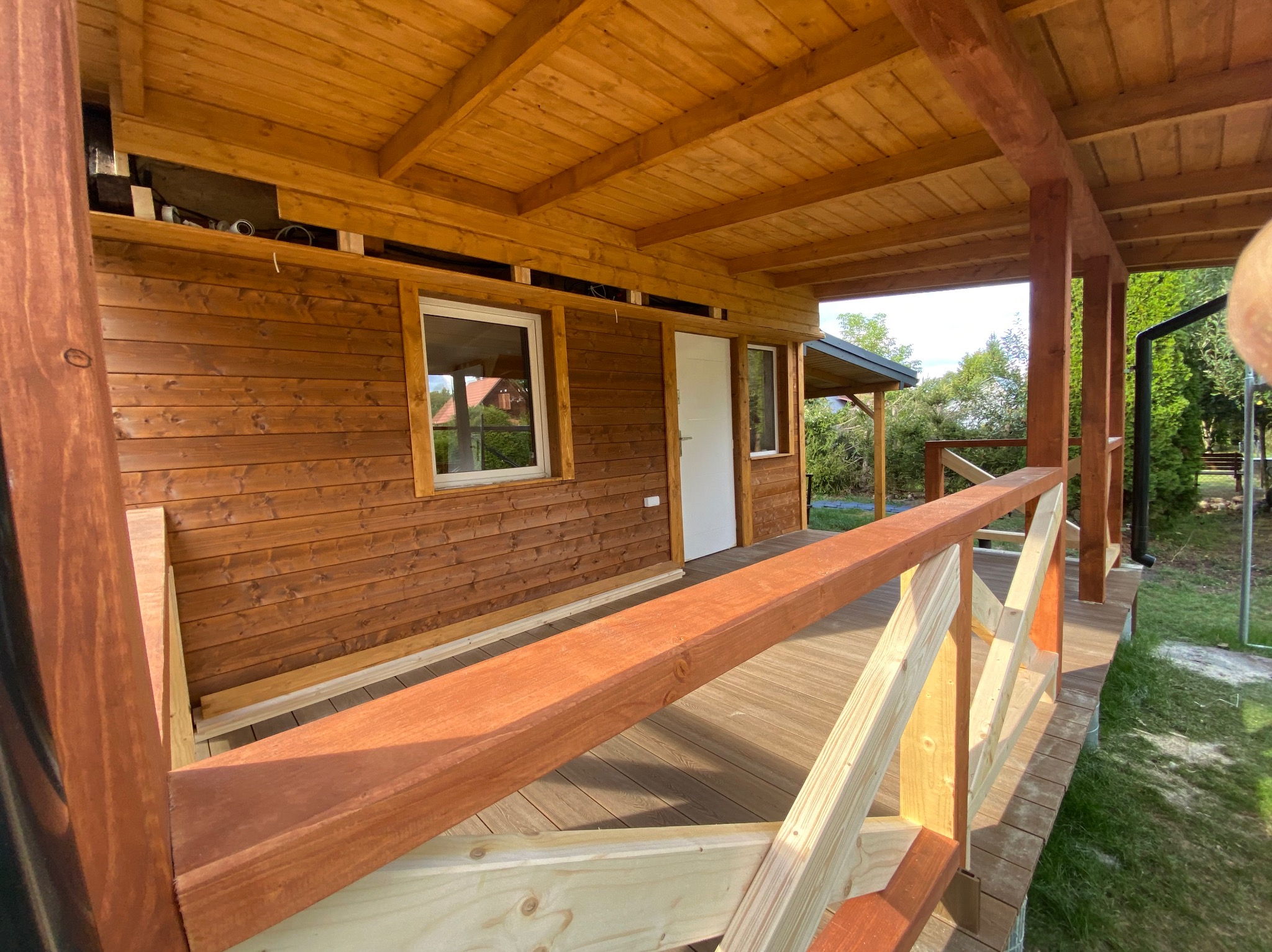 Zadaszony taras z drewnianą konstrukcją, widoczne deski podłogowe, balustrada w trakcie montażu z elementami surowego drewna i pomalowanymi na brązowo, drewniana elewacja budynku w tle.