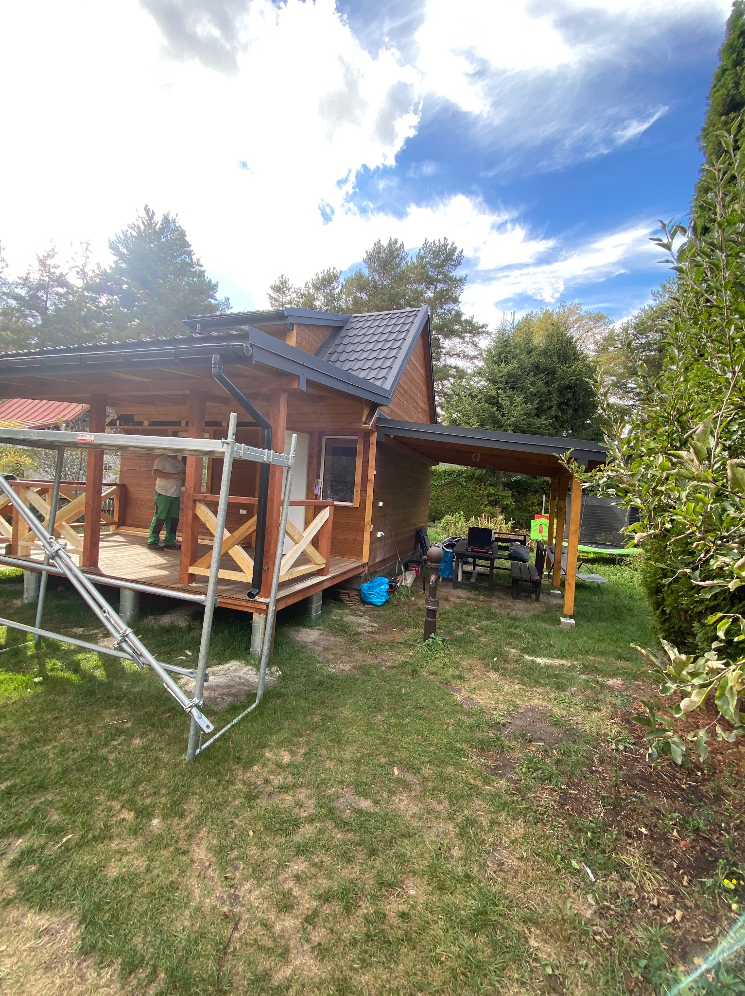Drewniany domek letniskowy z zadaszonym tarasem w trakcie budowy, widoczne rusztowanie i pracownik w zielonych spodniach, trawnik i drzewa w tle.