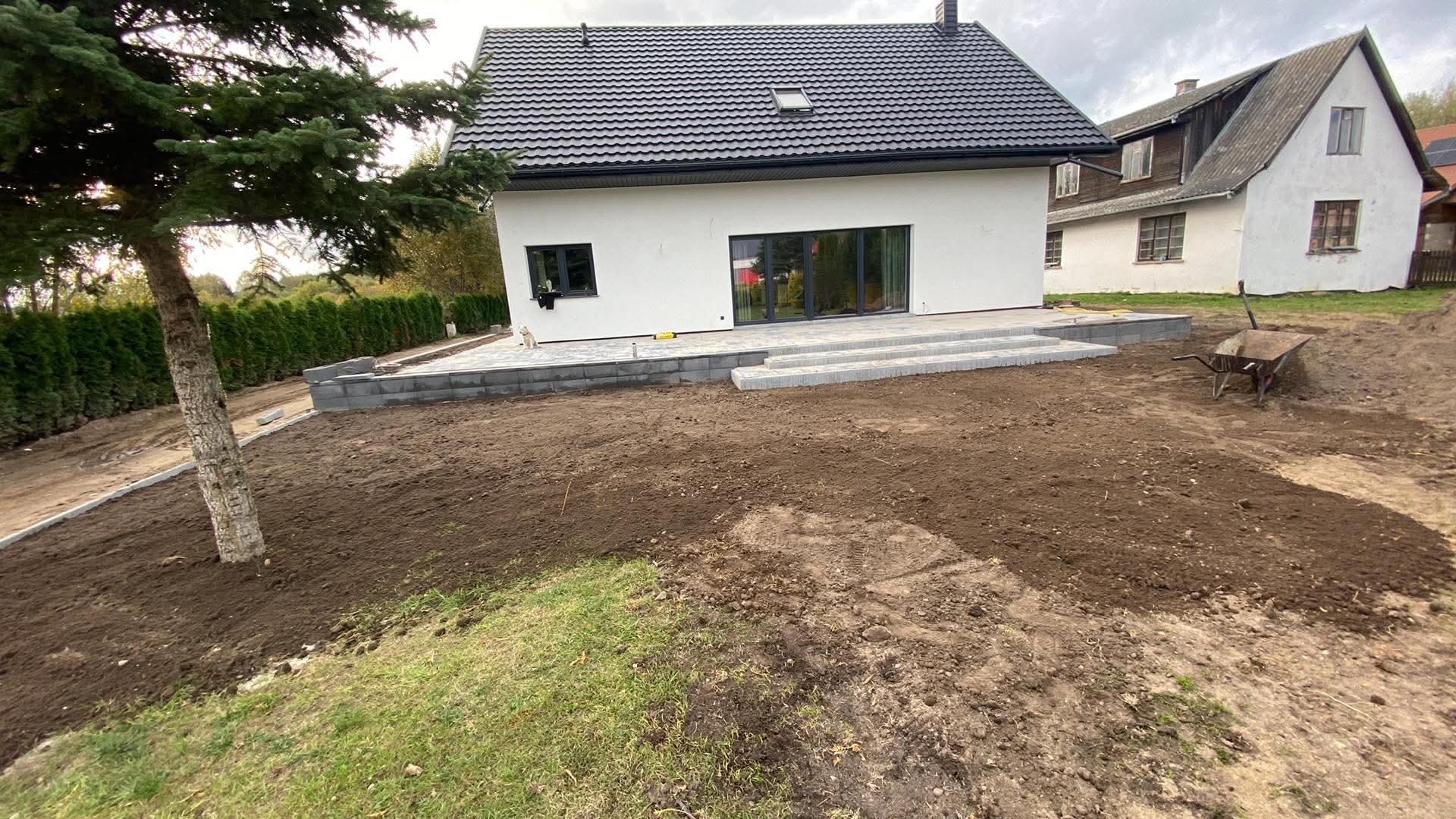 Nowoczesny dom z białymi ścianami i antracytowym dachem w trakcie budowy tarasu z kostki brukowej, widoczna taczka i przygotowany teren pod trawnik.