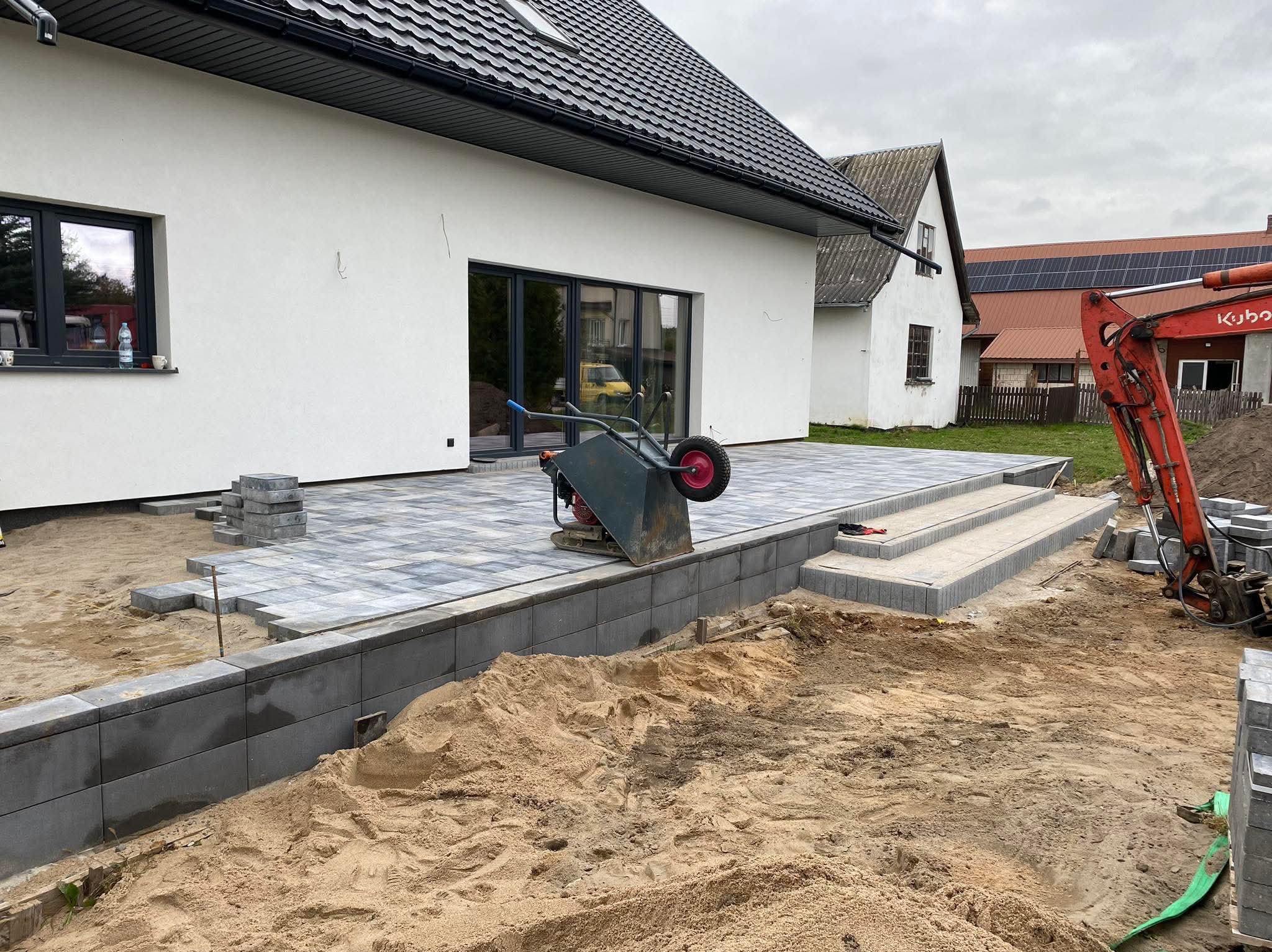 Układanie szarej kostki brukowej na tarasie przydomowym, z widoczną ubijarką, paletą kostki i mini koparką w tle.