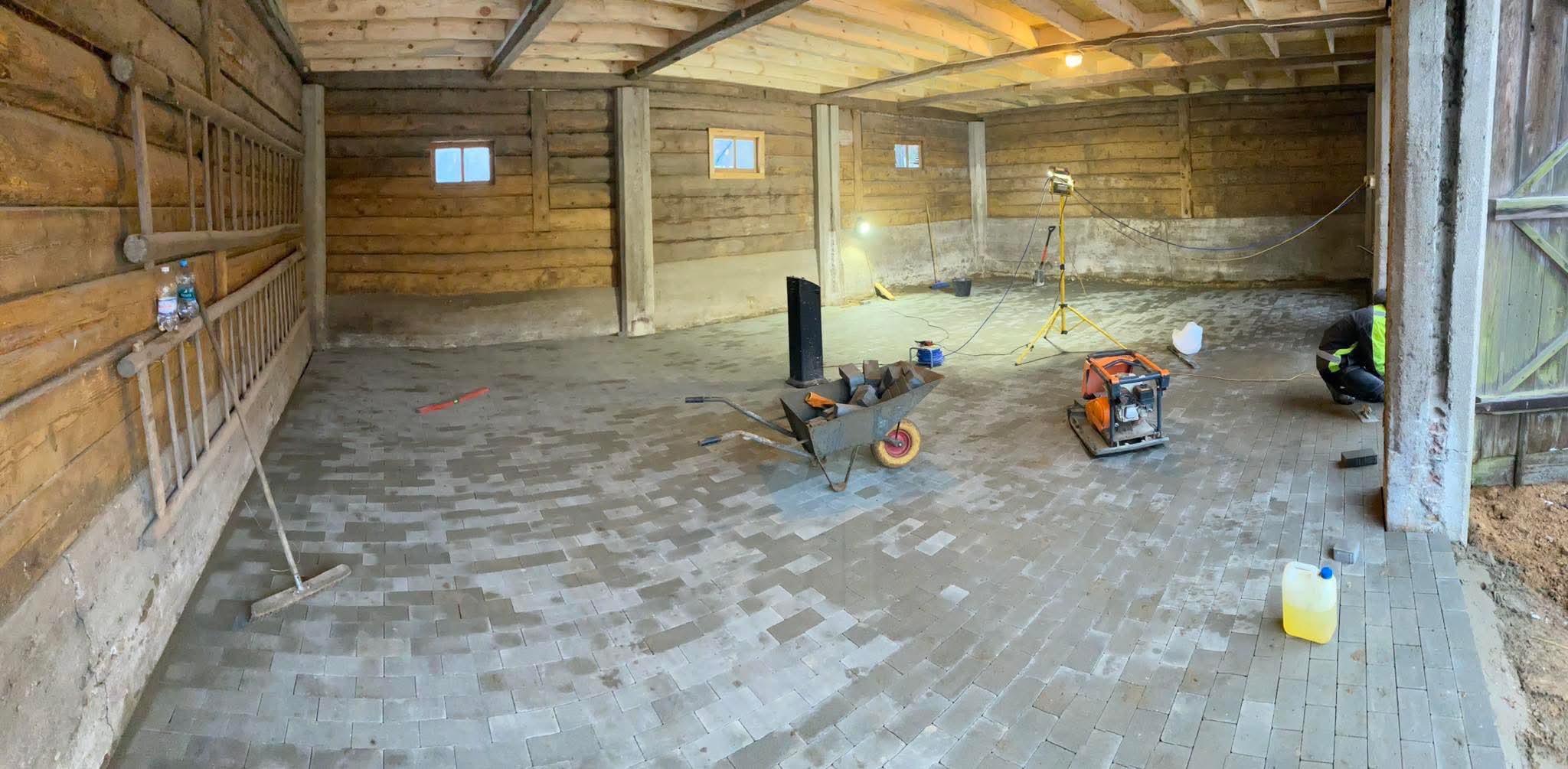 Układanie szarej kostki brukowej wewnątrz drewnianego budynku gospodarczego, widoczna taczka z kostką, ubijarka, oświetlenie budowlane i pracownik w odblaskowej kamizelce.