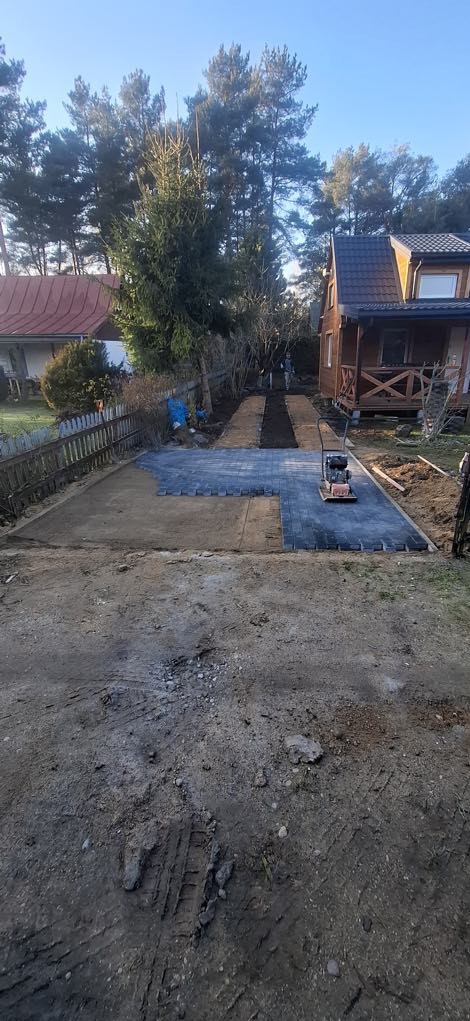 Układanie ciemnoszarej kostki brukowej na posesji, widoczna ubijarka wibracyjna, w tle dom z drewnianą werandą i drzewa.