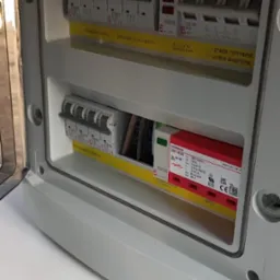 Otwarta skrzynka elektryczna z bezpiecznikami, z widocznymi zabezpieczeniami przeciwprzepięciowymi i oznaczeniami stanu pracy (normalna/awaryjna) na żółtych etykietach.