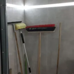 Garaż w Sosnowcu z zamontowanym oświetleniem LED podsufitowym i częściowo otwartą bramą garażową, w którym oparto o ścianę różnego rodzaju szczotki, kije i narzędzia ogrodowe.