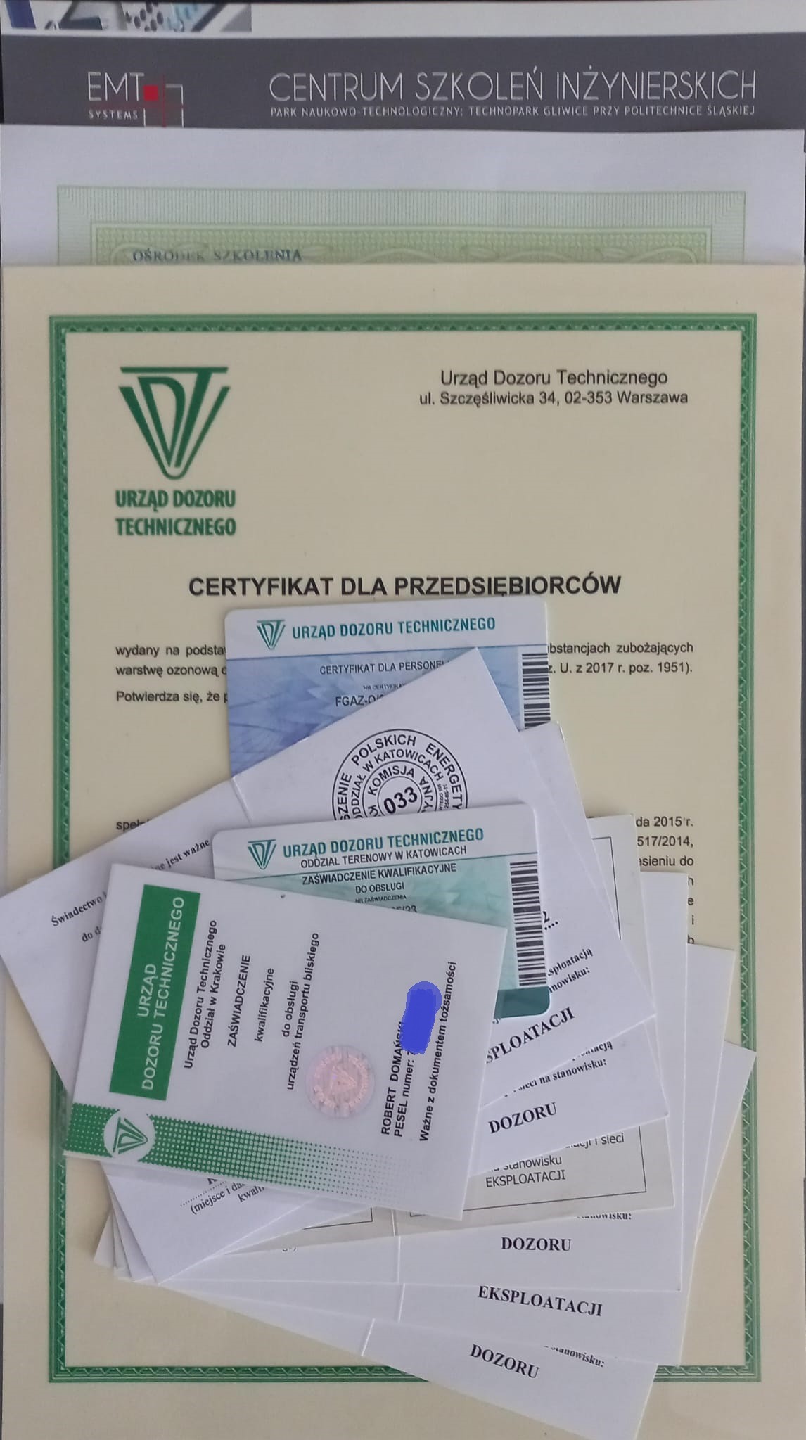Stos certyfikatów i zaświadczeń Urzędu Dozoru Technicznego, w tym certyfikat dla przedsiębiorców, zaświadczenie kwalifikacyjne i certyfikat dla personelu, częściowo zasłonięte, z widocznym logo EMT...