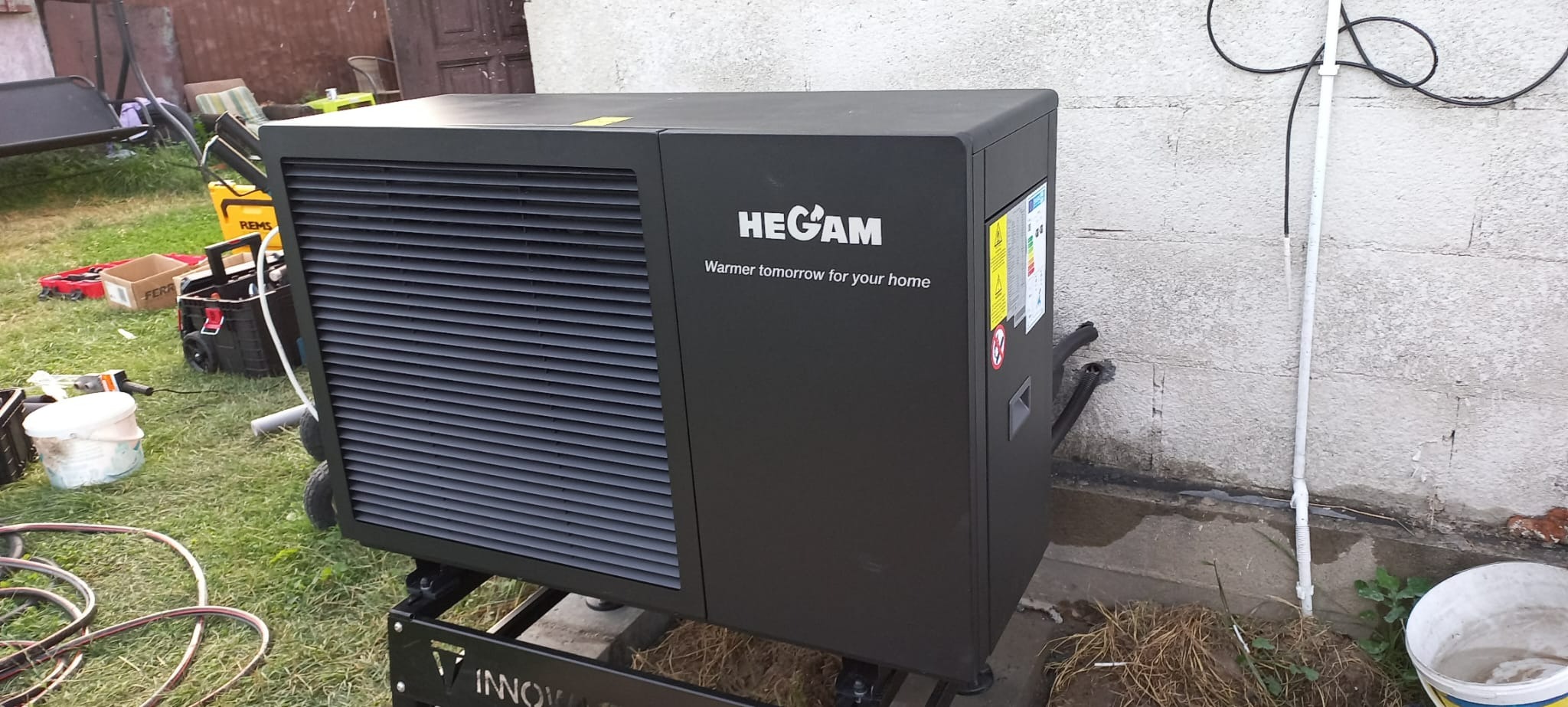 Czarna pompa ciepła HEGAM na trawie, widoczny kosz z narzędziami i skrzynka narzędziowa w tle, przy szarej ścianie budynku.