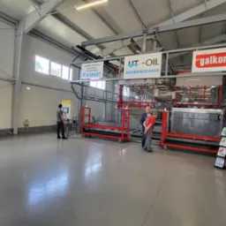 Wnętrze hali produkcyjnej z linią do galwanizacji, widoczni pracownicy obsługujący maszynę, pojemniki z chemikaliami oraz materiały reklamowe firmy.