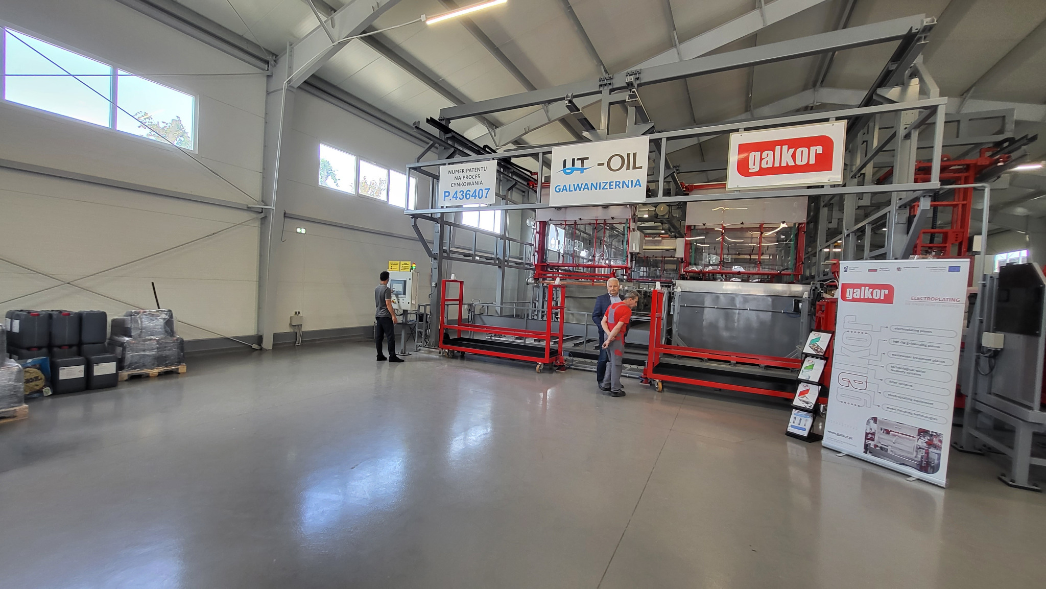 Wnętrze hali produkcyjnej z linią do galwanizacji, widoczni pracownicy obsługujący maszynę, pojemniki z chemikaliami oraz materiały reklamowe firmy.
