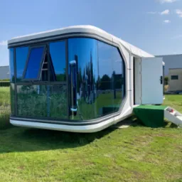 Nowoczesny, mobilny modułowy dom z dużymi oknami, otwartymi drzwiami i schodami, ustawiony na trawie. W oknach odbija się otoczenie. W tle budynki i osoba na kosiarce.