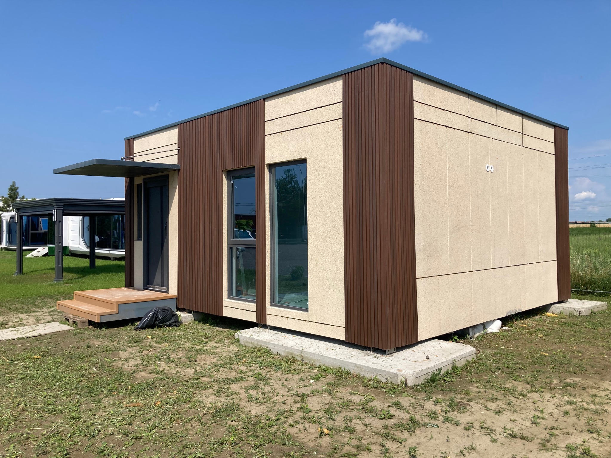Nowoczesny, parterowy dom modułowy z brązowymi panelami i drewnianymi schodami wejściowymi, osadzony na betonowych fundamentach na tle zielonego pola i błękitnego nieba.