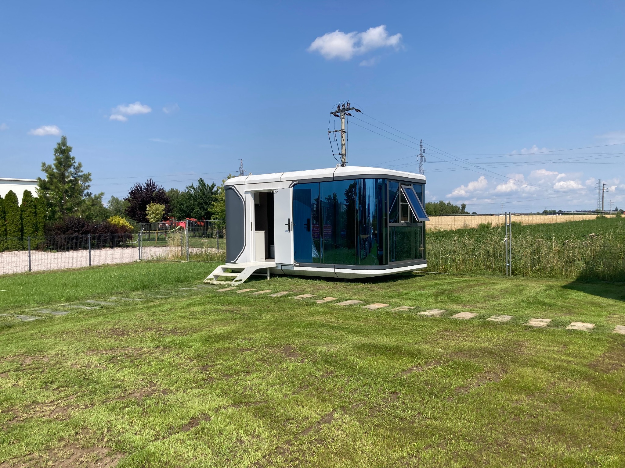 Nowoczesny, przeszklony modułowy dom na zielonej trawie w słoneczny dzień. Otwarta witryna okienna, schody wejściowe. W tle pola i słupy energetyczne.