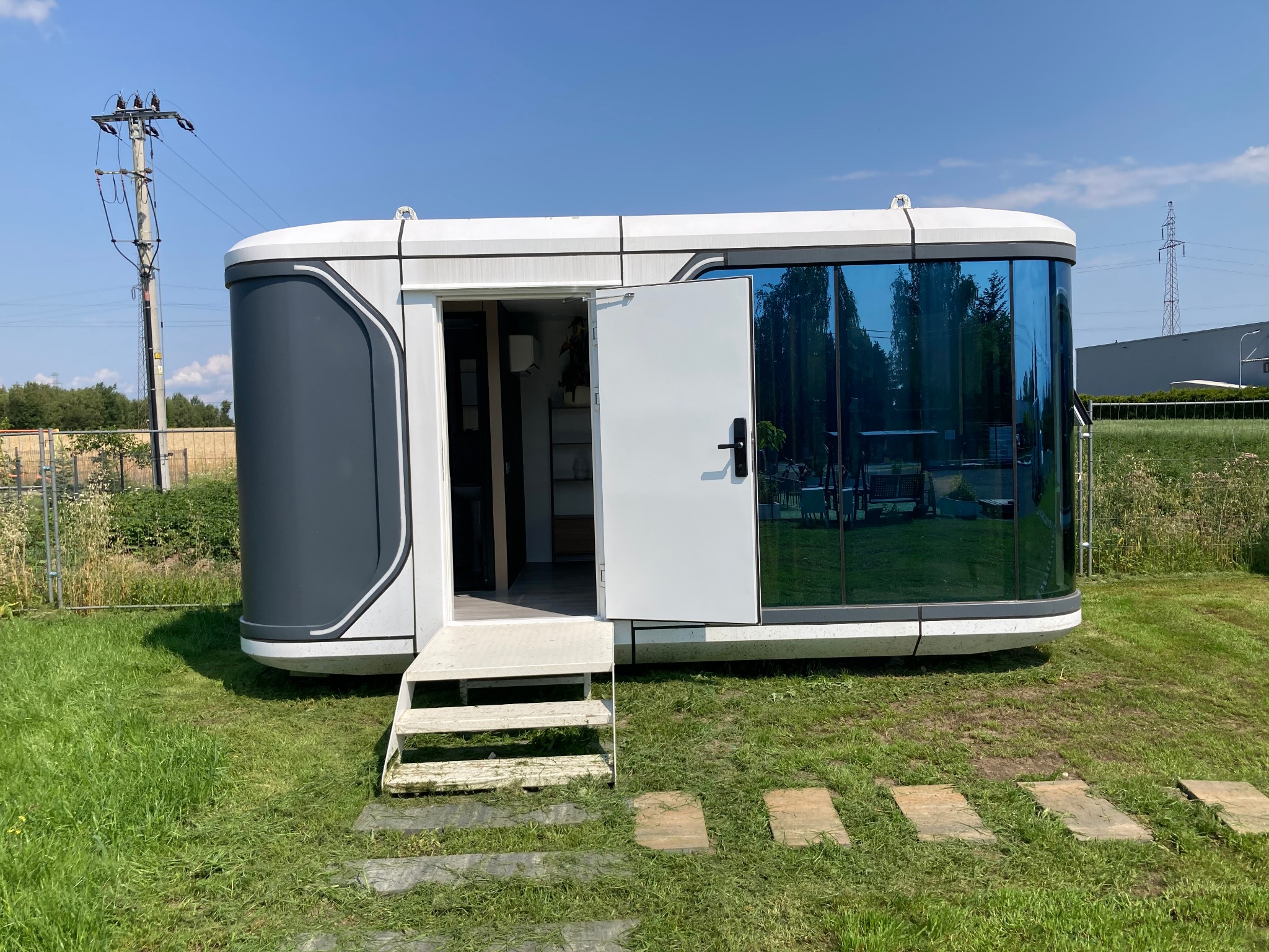 Nowoczesny, mobilny dom modułowy z otwartymi drzwiami i schodami na trawniku, z refleksami drzew w niebieskich szybach. Widoczny słup energetyczny w tle.