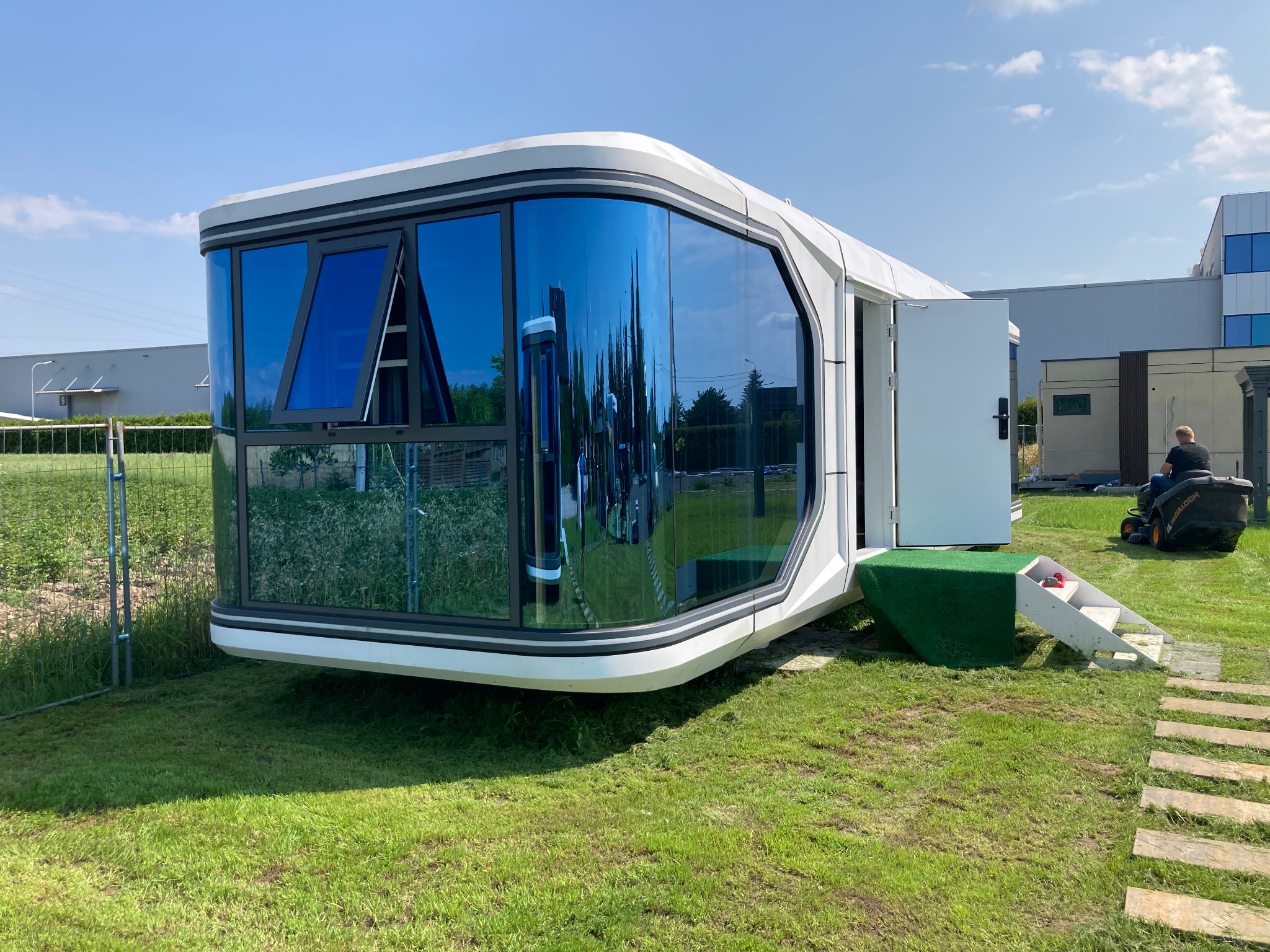 Nowoczesny, mobilny modułowy dom z dużymi oknami, otwartymi drzwiami i schodami, ustawiony na trawie. W oknach odbija się otoczenie. W tle budynki i osoba na kosiarce.