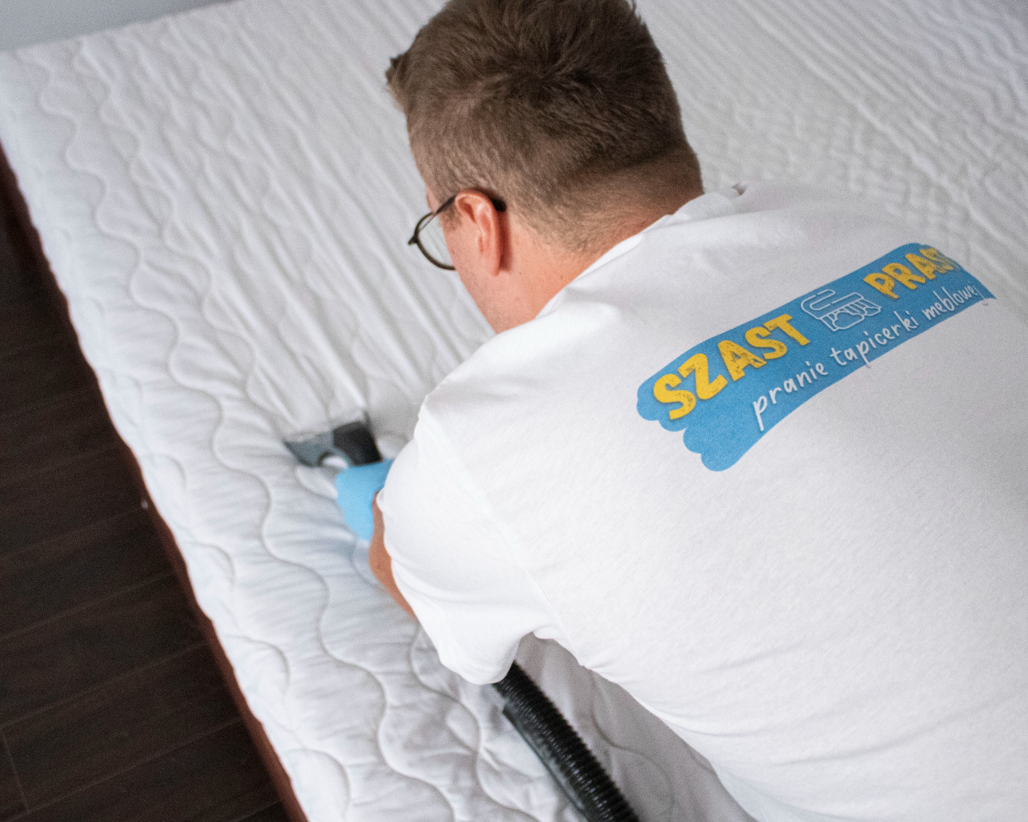 Mężczyzna w okularach, w koszulce z logo firmy czyszczącej tapicerkę meblową, czyści biały materac za pomocą odkurzacza piorącego.