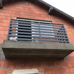 Szara, pozioma balustrada balkonowa z płaskowników, zamontowana na betonowej płycie balkonu w surowym stanie, na tle ściany z czerwonej cegły.