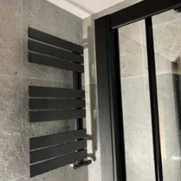 Czarny grzejnik drabinkowy na szarej, kwadratowej ścianie z płytek, obok czarnej ramy kabiny prysznicowej z przezroczystym szkłem.