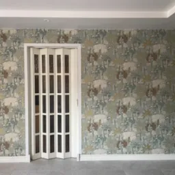 Pokój z tapetą w motywy roślinne i składanymi, białymi drzwiami, widoczne szare płytki podłogowe i białe listwy przypodłogowe.