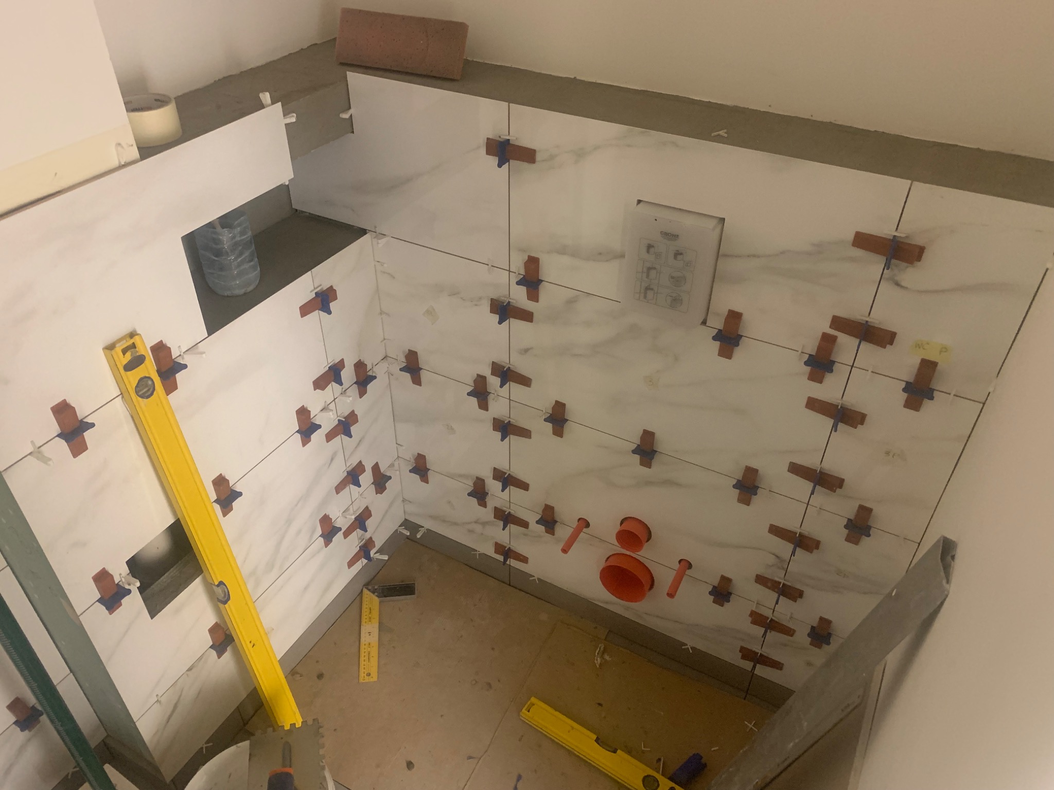 Białe płytki imitujące marmur, układane na ścianie w łazience z użyciem krzyżyków dystansowych, widoczne poziomice i rury kanalizacyjne.