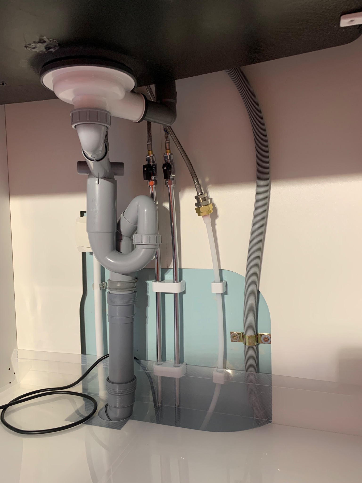 Szare rury odpływowe i dopływowe pod zlewem w szafce, widoczne połączenia i wężyki, przewód elektryczny na dnie szafki.