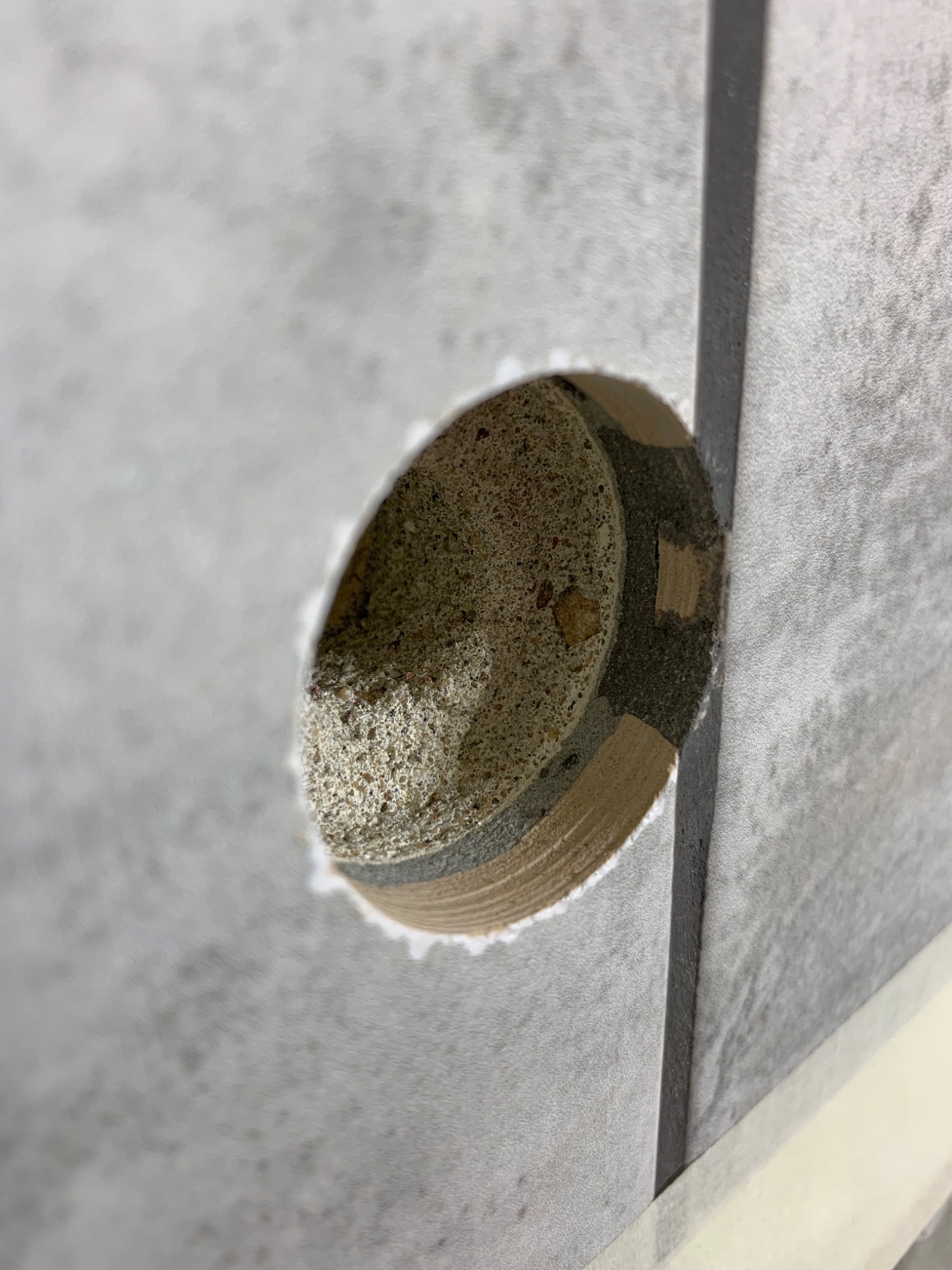 Wywiercony otwór w szarej płytce ściennej, widoczny beton i fragmenty drewnianych kołków mocujących, zabezpieczone taśmą malarską.