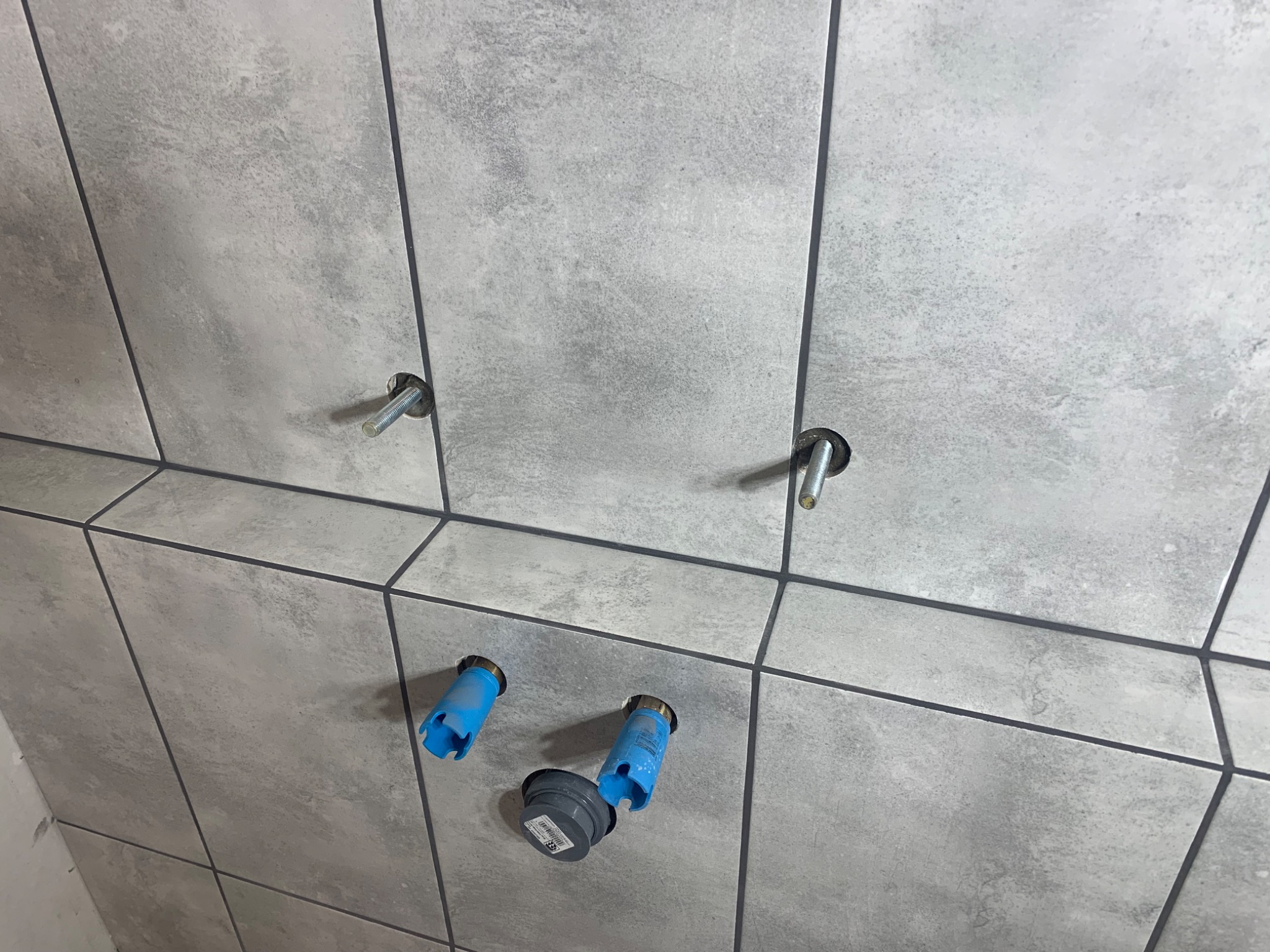 Szare płytki ceramiczne na ścianie z przygotowanymi otworami na armaturę, widoczne śruby mocujące i plastikowe osłonki na rury.