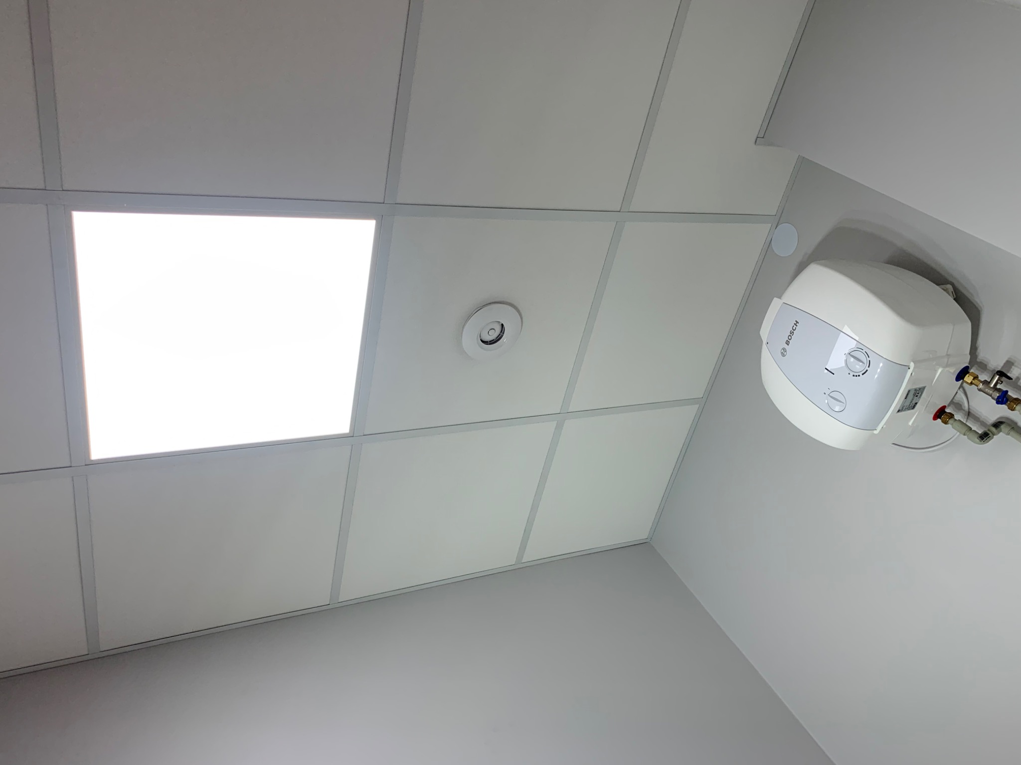 Biały sufit podwieszany kasetonowy z wbudowanym panelem LED, widoczny okrągły czujnik oraz biały bojler elektryczny marki Bosch z podłączonymi rurami.