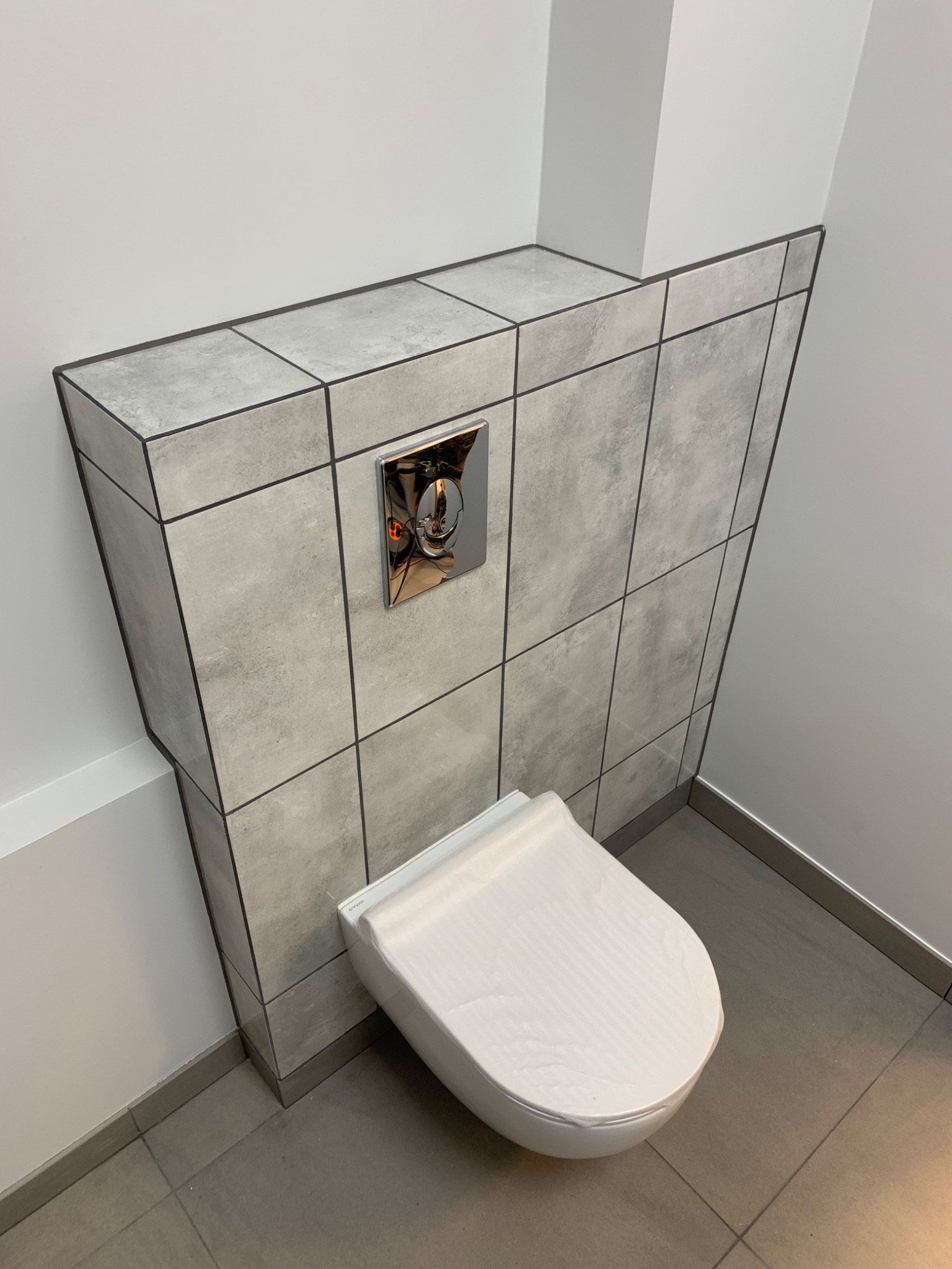 Nowoczesna toaleta podwieszana, zabezpieczona folią, z chromowanym przyciskiem spłukującym, obudowana płytkami ceramicznymi w odcieniach szarości z ciemnymi fugami.