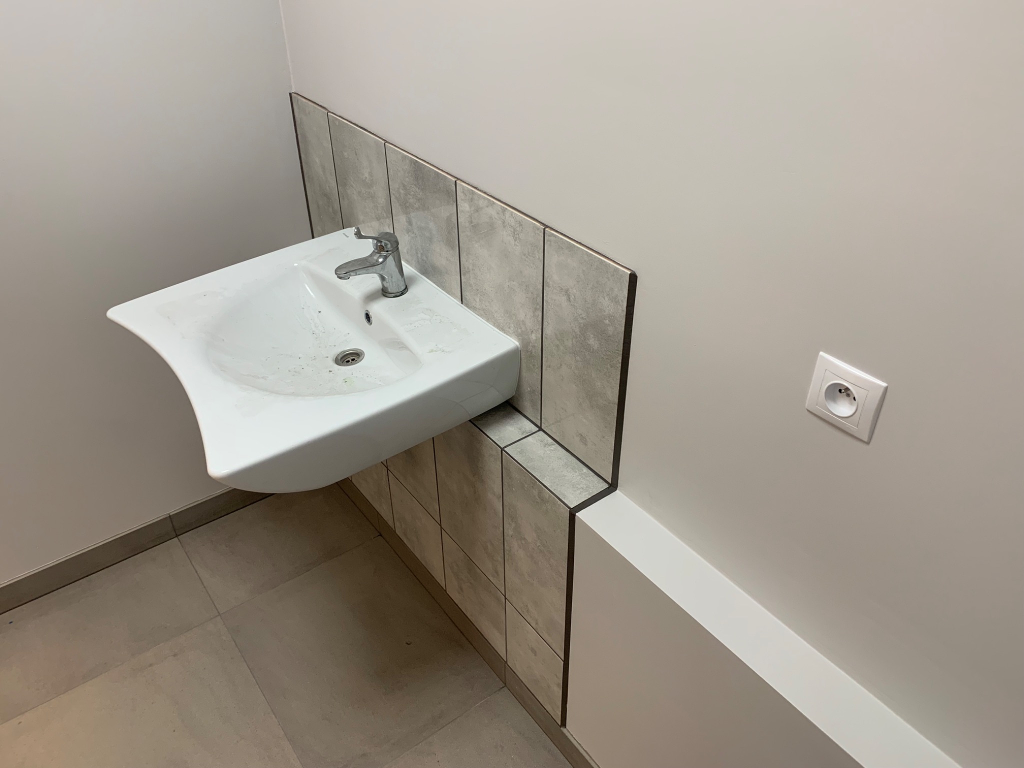 Biała umywalka zamontowana na ścianie z szarymi płytkami, widoczne połączenie płytek i biała listwa przypodłogowa, obok gniazdko elektryczne.