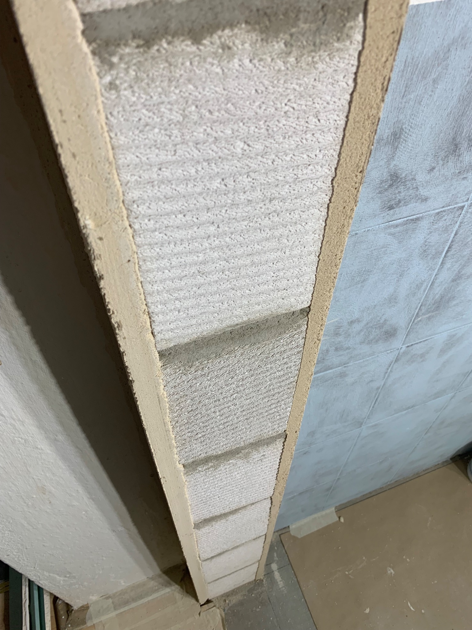 Wąski fragment ściany z płyt kartonowo-gipsowych, widoczne łączenia i szpachlowanie, sąsiadujący z wykończoną ścianą z jasnoniebieskich płytek.